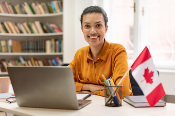 Online foreign languages tutoring. Happy female teacher sitting in library with flag of Canada, using laptop