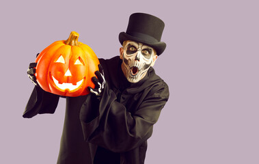 Man in Halloween costume. Skeleton in black cloak and top hat standing isolated on light purple background, holding orange jack o lantern and looking at camera with funny surprised face expression