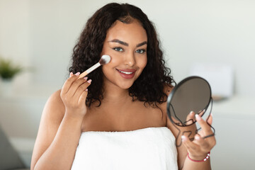 Daily makeup. Beautiful oversize black lady applying blush with makeup brush, holding mirror and...