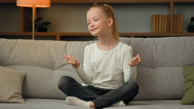 Funny active little girl sit on couch in lotus pose snap fingers beat rhythm listening favorite pop music dancing move by loud musical track creative trendy baby style dance enjoying free time at home