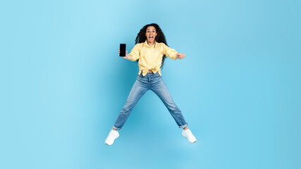 Excited Middle Eastern Woman Showing Phone Jumping Over Blue Background