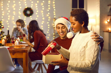 Merry Christmas my love. Man surprises his beloved woman by giving her present during family Christmas dinner. African American woman opens gift box sitting next to Caucasian man. Blurred background.