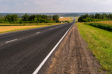 An empty roadway outside the city passes by a rural field and a blooming meadow. Travel by car away from the city and the hustle and bustle. Paved road on a sunny day without cars.