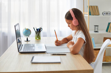 School child studying online at home. Profile view little girl in pink headset sitting at desk in...