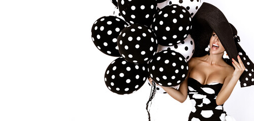 Beautiful fashion model, dressed in pinup style black dress in white polka dot and elegant hat is holding balloons with dots on white background. Retro style. Polka dot fashion.