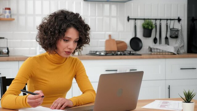 Smiling curly woman student study talking listens online video conference call webcam laptop, writes in notebook. Positive businesswoman communicates remote connection chat computer at home kitchen