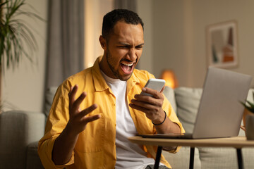 Work stress concept. Angry young black man shouting into smartphone, working online on laptop pc at home office