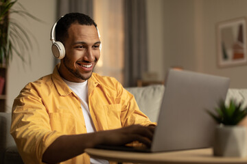 Joyful young African American man using laptop, having remote job, wearing headphones at home