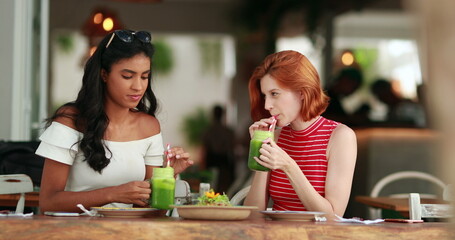 Two diverse pretty girls drinking green juice. Multicultural young women holding healthy veggie juice