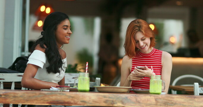Two multiracial friends at cafe showing cellphone screen smiling and feeling happy