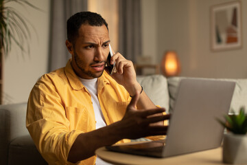 Fototapeta na wymiar Upset millennial black man calling on smartphone, gesticulating, looking at laptop screen in home office
