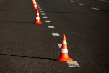 A row of traffic orange striped cones on the road. These objects are temporary traffic control devices for directing and avoiding sections of the road being repaired or diverting traffic.