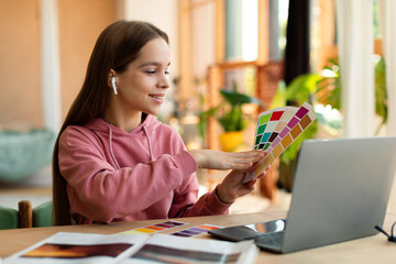 Happy smart teen girl having online design courses with teacher via laptop computer, holding color palettes