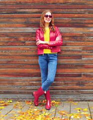 Portrait of happy smiling young woman wearing red leather jacket, heart shaped sunglasses in the city on wooden wall background