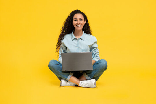 Happy young woman using laptop pc for online work or studies, sitting cross legged on yellow studio background