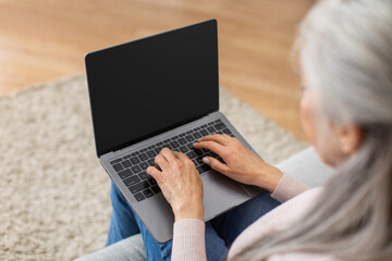 Cropped caucasian elderly gray-haired woman typing on laptop with blank screen in living room