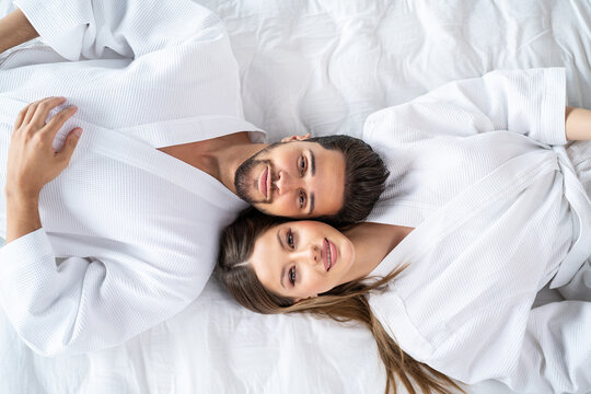 Young happy couple relaxing, lying in bed in white bathrobes, head to head, looking at camera and smiling.