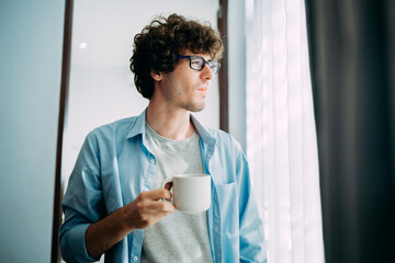 Portrait of a young handsome caucasian with a cup of coffee looking outside at window in his home...