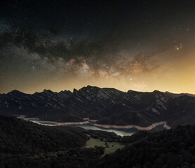 Milky way with stars rising over a beautiful mountain range, water reservoir and forest. Long exposure night photo and astrophotography in nature landscape.