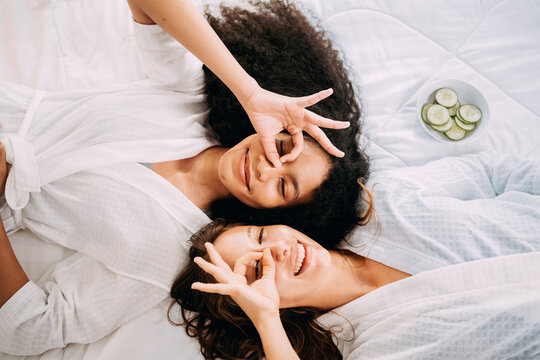 Happy young women in white bathrobe towels apply slice cucumber facial masks together lying on the bed, LGBTQ couples fun leisure activities spa beauty natural treatment skin care party.