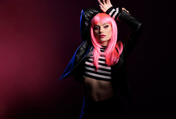 Portrait of sensual beauty model with pink hair wig and rocker jacket standing over background with dark shadows and light. Sexy stylish woman with punk trendy fashion items doing pose.