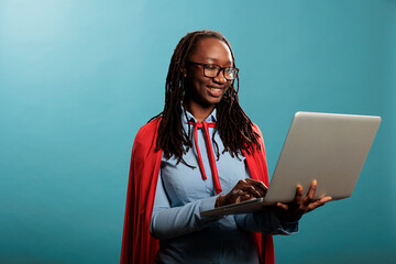 Brave and proud young adult superhero woman using modern laptop while standing on blue background....