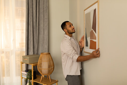 Young black man putting picture frame, hanging painting on wall, empty space. Home interior and domestic decor