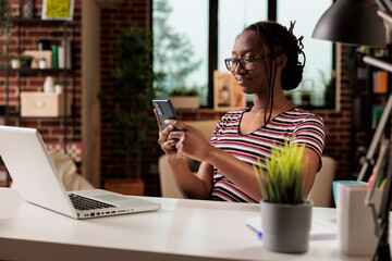 Smiling woman messaging on smartphone, chatting in social network at home office desk. Employee in glasses having break from work, remote student surfing internet on mobile phone