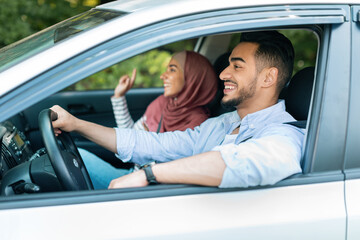 Glad smiling millennial islamic husband driving at steering wheel with wife in hijab, ride in car, enjoy music