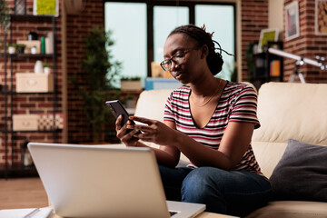 Young african american freelancer holding smartphone, typing message, chatting in social network. Remote worker having break, surfing internet on mobile phone, sitting on couch