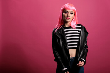 Young adult with pink hair wearing biker leather jacket in studio, posing with attractive rocker style in front of camera. Sensual carefree woman with elegant punk clothes to feel trendy and pretty.
