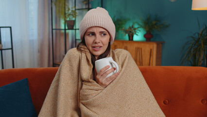 Sick girl wear hat wrapped in plaid sit alone shivering from cold on sofa drinking hot tea in...
