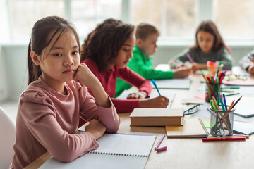 Unhappy Asian Schoolgirl Thinking Sitting With Diverse Classmates In Classroom