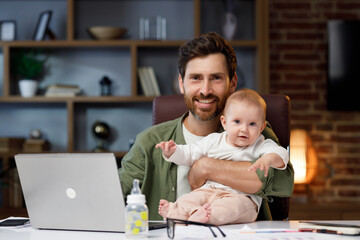 Portrait of a Father on maternity leave, in his home office with a small child in his arms. Dad at...