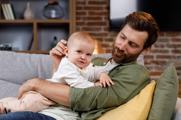 Dad plays with his little baby, on the couch in a cozy apartment. Fatherly love. Father hugs and kisses his little daughter.