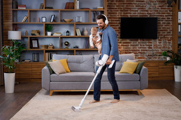 A young father, with a child, does housework. Father on maternity leave. Dad cheerfully sings, dances and vacuums, cleans with a small child in his arms. Paternity Leave