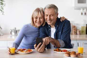 Positive elderly husband and wife using smartphone while having breakfast