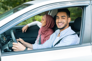 Satisfied millennial arab male driving, looks at camera, rides with his wife in hijab in car and enjoys journey
