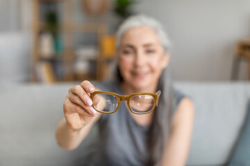 Cheerful caucasian elderly gray-haired female holding glasses in living room interior, selective focus, blurry