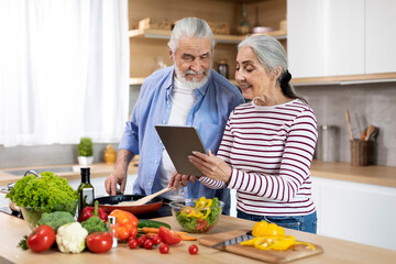 Happy Elderly Spouses Cooking Food In Kitchen And Using Digital Tablet Together