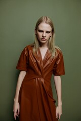 Confidence young woman looks at camera with expressive glade evening smoky eyes makeup dress in brown leather trendy trench coat posing isolated at studio olive green wall background. Resale offer