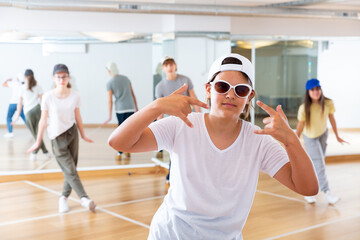 Fototapeta na wymiar Portrait of teen girl in cap and sunglasses performing hip-hop with friends at group dance class