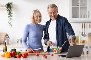 Loving senior spouses cooking together at home, using laptop