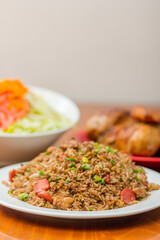 Chaufa rice in a white plate, on a wooden table, with an out of focus background grilled chicken and salad.