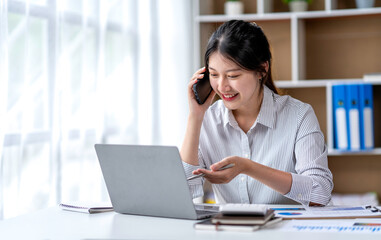 Fototapeta premium Young woman talking on the phone working on laptop in office. Asian businesswoman sitting in her office in office. Beautiful woman freelancer working online at her home.