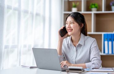 Fototapeta premium Young woman talking on the phone working on laptop in office. Asian businesswoman sitting in her office in office. Beautiful woman freelancer working online at her home.