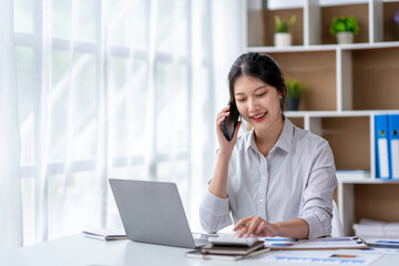 Young woman talking on the phone working on laptop in office. Asian businesswoman sitting in her office in office. Beautiful woman freelancer working online at her home.