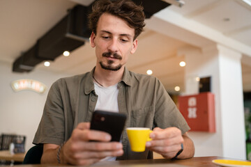 Fototapeta na wymiar one caucasian man young adult millennial male using mobile phone sit at table in cafe at the table alone happy smile confident having online chat using internet for text messages or social network