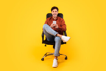 Happy Man Using Cellphone Sitting In Office Chair, Yellow Background