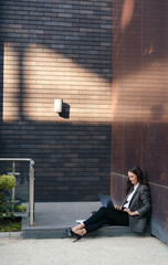 Attractive modern lady sitting with crossed legs on the threshold of the office building and using...